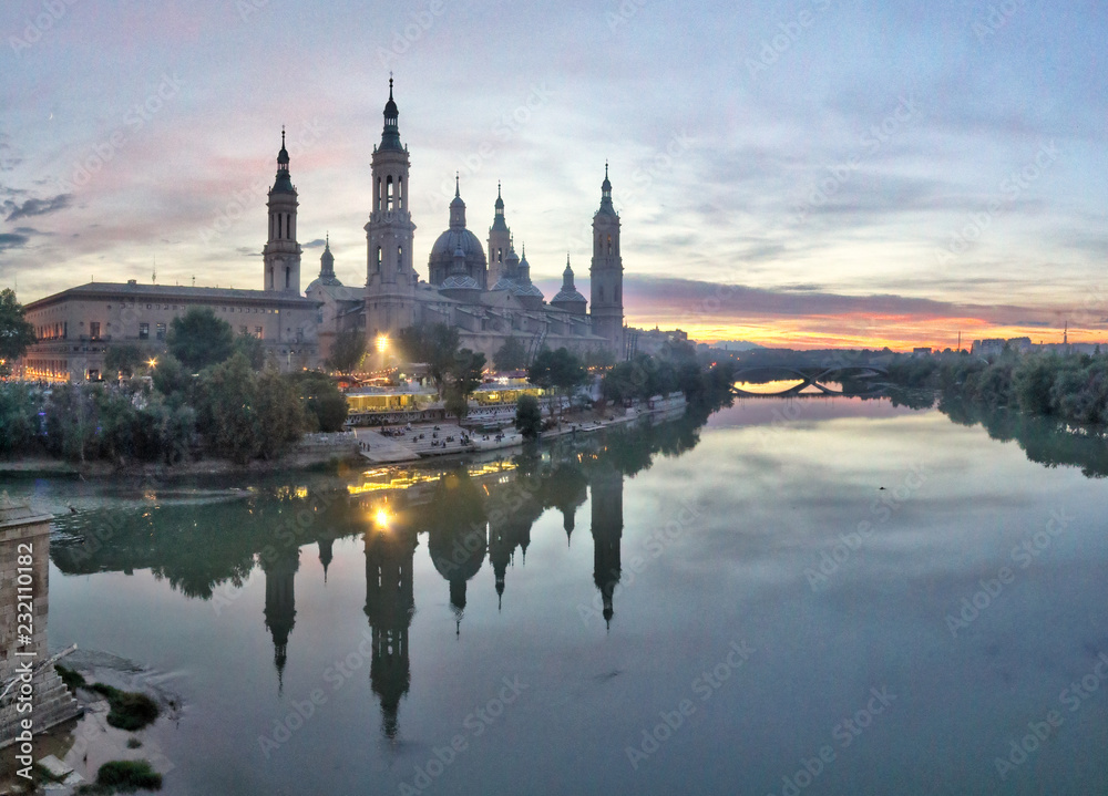A landscape of Pilar Cathedral and Santiago Bridge reflecting in the Ebro river at sunset, after a storm, in a cloudy autumn, in Zaragoza, Spain