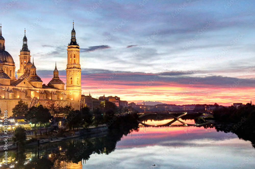 Naklejka premium A landscape of Pilar Cathedral and Santiago Bridge reflecting in the Ebro river at sunset, after a storm, in a cloudy autumn, in Zaragoza, Spain