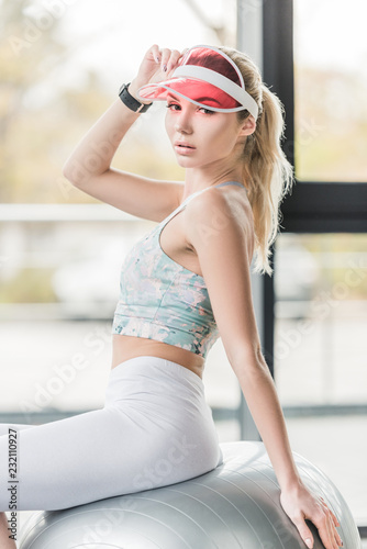 stylish sportswoman in visor hat looking at camera and posing on grey fitness ball at gym