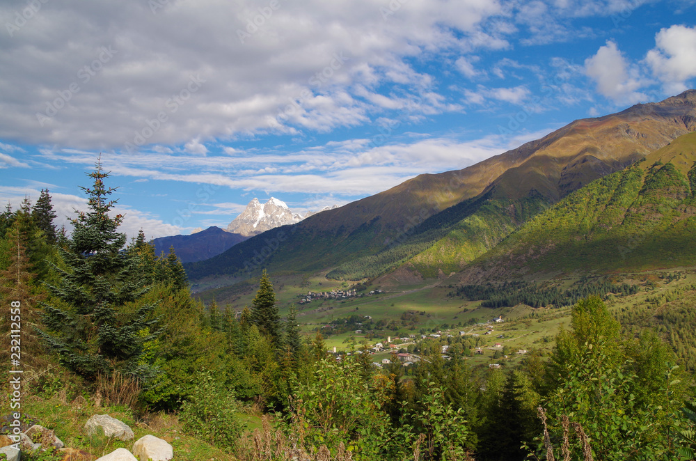 Scenic autumn landscape in the mountains of the Caucasus. View of the valley and village on the background of snowcapped peak of the Ushba Mountain. Nature and travel. Georgia, Svaneti region