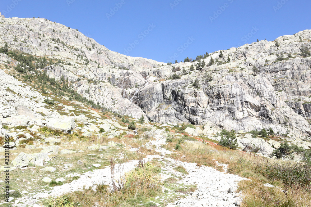 A path going through high rocky mountains with firs and pine trees forest and a blue sky in a sunny autumn, in Panticosa, Aragon Pyrenees, Spain