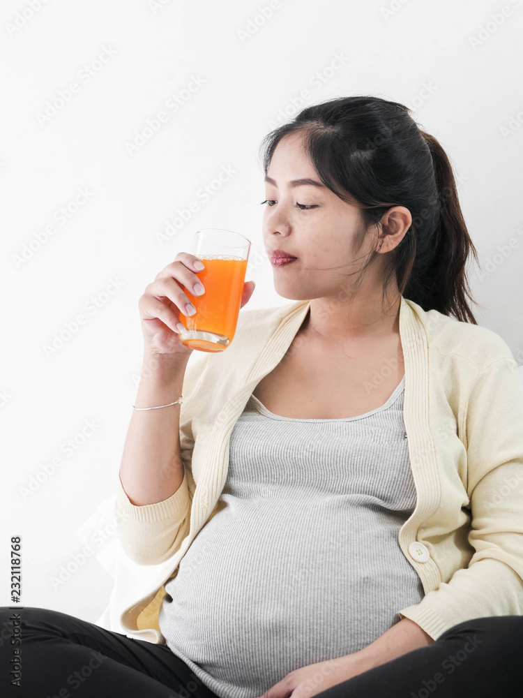 Asian pregnant woman holding a glass of orange juice and sitting on bed, lifestyle concept.