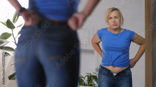 Beautiful adult blonde woman struggling to fit into old too tight pair of jeans after gaining weight while standing in front of mirror at home. Reflection of sad female trying to squeeze into pants.