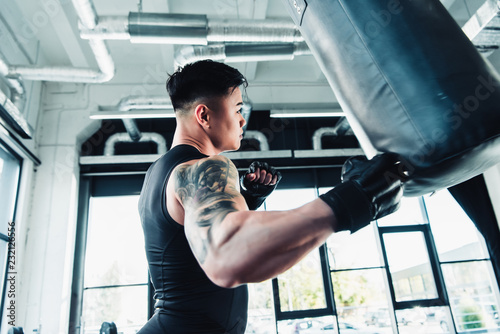 side view of young asian sportsman wearing boxing gloves and punching boxing bag at gym