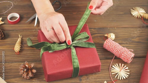 Close-up top view of female hands decorating red gift boxes with backer s twine and fir brunches on wooden table. Pine cones and christmas decoration on dark desk. photo