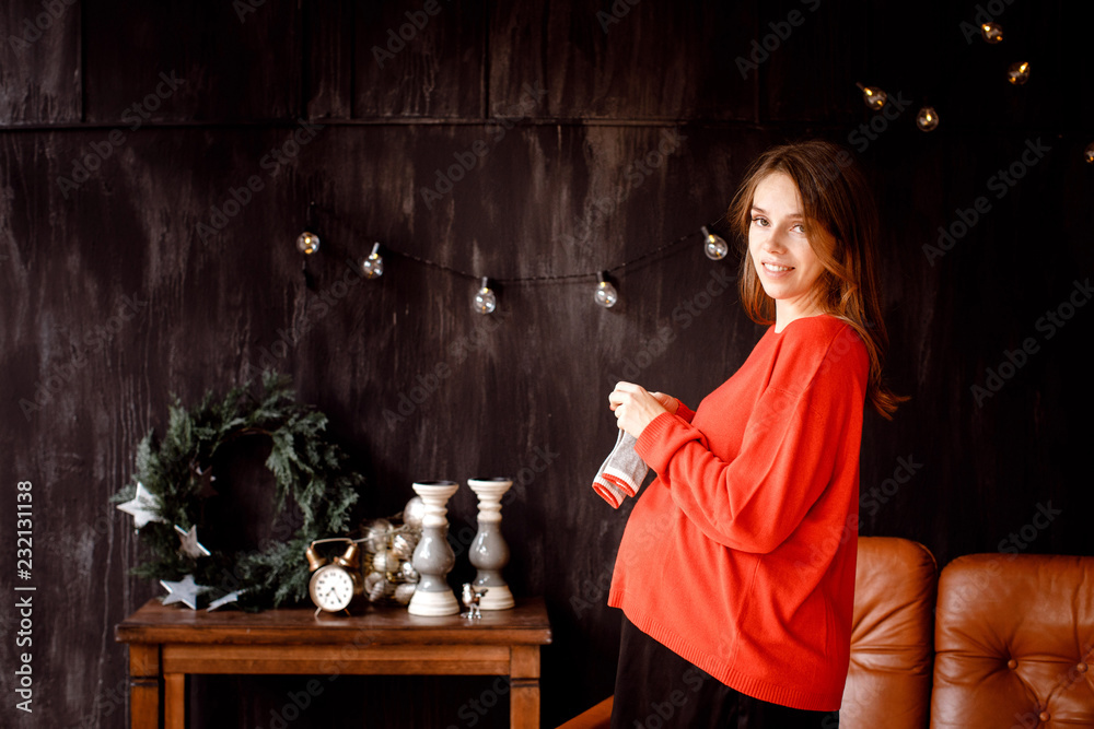 Side view of young pregnant female smiling and looking at camera while holding baby socks and standing in stylish room decorated for Christmas Side view of young pregnant female smiling and looking at camera while holding baby socks and standing in stylish room decorated for Christmas