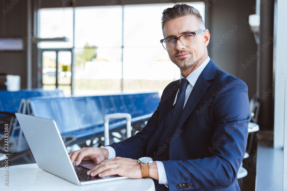 handsome businessman in glasses looking at camera and using laptop in airport handsome businessman in glasses looking at camera and using laptop in airport