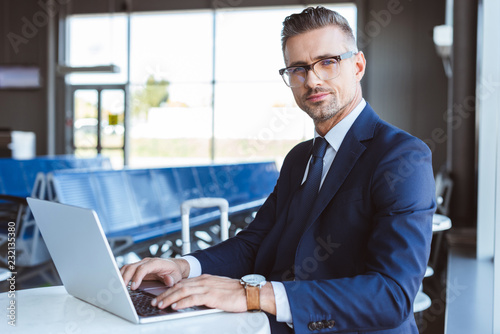 handsome businessman in glasses looking at camera and using laptop in airport