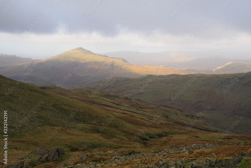 Obraz premium Sunlight falls on a Lake District mountain top out of an overcast and cloudy sky lighting it up with warm Autumn colours, Cumbria, UK