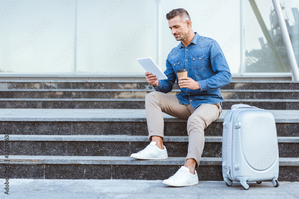 Handsome adult man with a coffee to go sitting on the staircase with luggage and using digital tablet Handsome adult man with a coffee to go sitting on the staircase with luggage and using digital tablet