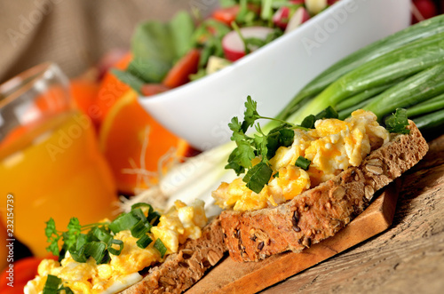 Close-up of wholemeal bread with scrambled eggs, fresh herbs, spring onions, tomatoes and salad bowl in background - healthy breakfast