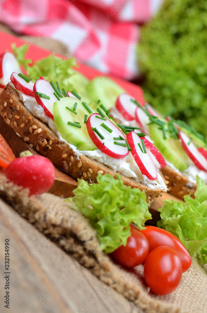 Close-up of wholemeal grain bread with curd cheese, fresh radish, cucumber and tomatoes on a wooden cutting board - concept of healthy fitness breakfast or snack, fresh salad in background