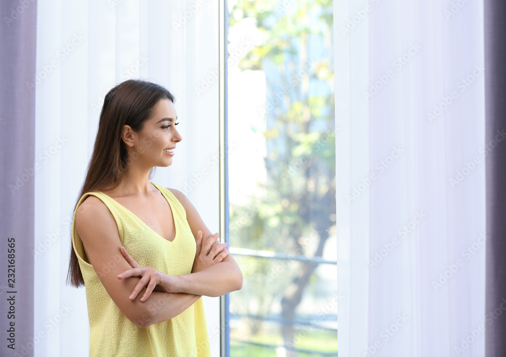 Young woman near window with open curtains at home. Space for text Young woman near window with open curtains at home. Space for text