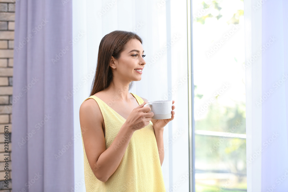 Young woman with cup of drink near window with open curtains at home Young woman with cup of drink near window with open curtains at home