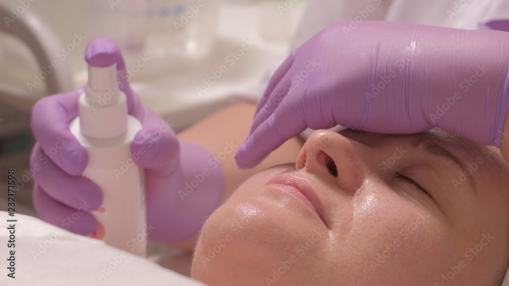 Naklejka premium A professional cosmetologist in lilac gloves with one hand covers his eyes, while the other sprays a moisturizing tonic on the face of a middle-aged woman.