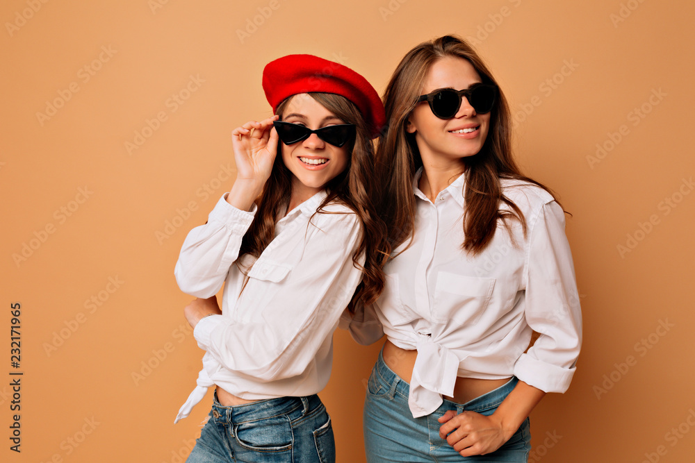Inside portrait of two stylish sisters posing at camera and smiling with true happy smile wearing bright pullovers over beige background wearing black sunglasses and white shirts Inside portrait of two stylish sisters posing at camera and smiling with true happy smile wearing bright pullovers over beige background wearing black sunglasses and white shirts
