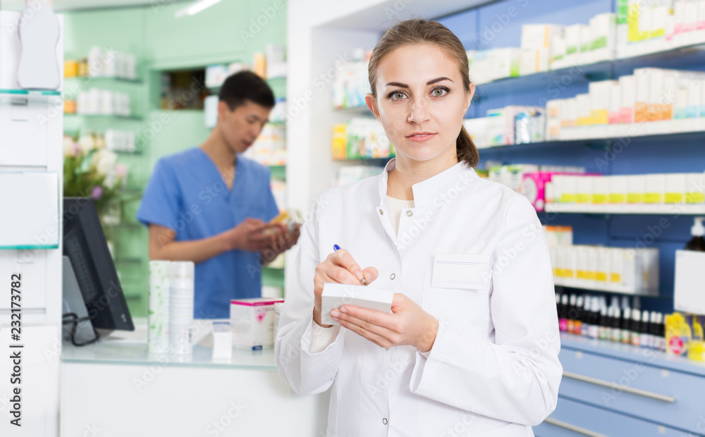 Young female pharmacist is attentively checking medicine to notebook