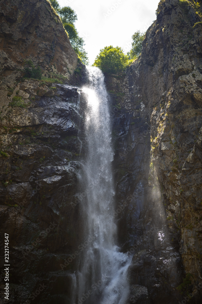 A high waterfall with white splashes of water descends from the mountain and sparkles in the sun. Attraction of Georgia Gveleti waterfall