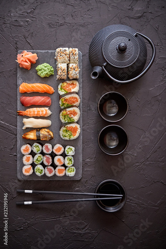 Assortment of different kinds of sushi rolls placed on black stone board. Traditional asian iron tea pot on side. Top angle view.