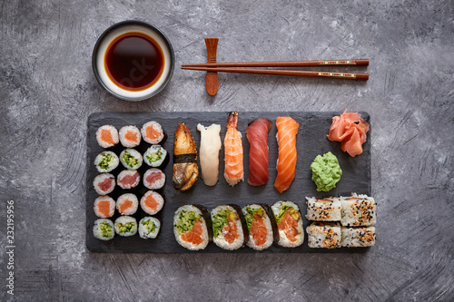 Composition of different kinds of sushi rolls placed on black stone board. Chopsticks and soy sauce bowl on side. Top angle view.