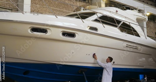 Young man (guy) specialist in white work coat polishes the yacht in the garage. Concept of: Preparation, Cleaning, Polishing, Master, Professional, After painting, Using chemistry.