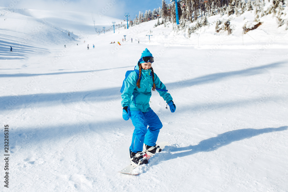A beautiful girl in winter clothes is riding a snowboard. She is wearing a blue helmet and a green jacket. having a great time in the mountains. Concept of travel, leisure, freedom, sport.