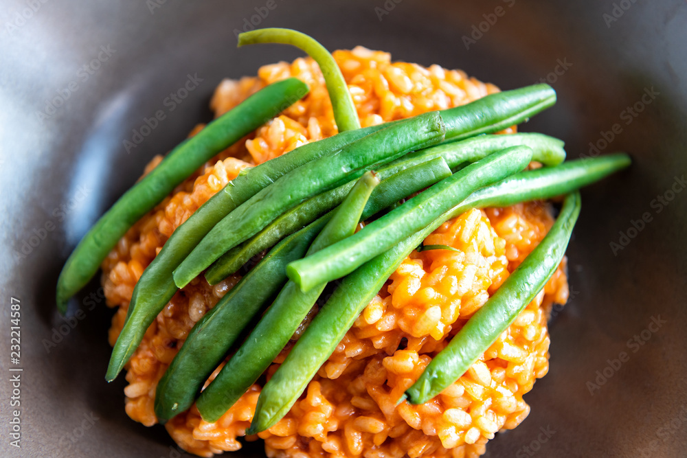 Macro closeup flat top view of risotto Italian arborio cooked rice with tomato sauce, olive oil, green beans on top in brown dish, plate on table Macro closeup flat top view of risotto Italian arborio cooked rice with tomato sauce, olive oil, green beans on top in brown dish, plate on table