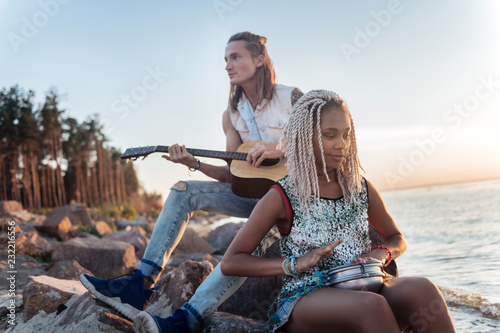 Sitting on rock. Blonde-haired athletic man playing the guitar while sitting on rock near beach