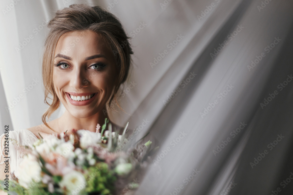 Gorgeous beautiful bride with bouquet posing at window in soft light. Happy Bride portrait in stylish gown, with perfect makeup, smiling and dreaming. Romantic moment. Space for text Gorgeous beautiful bride with bouquet posing at window in soft light. Happy Bride portrait in stylish gown, with perfect makeup, smiling and dreaming. Romantic moment. Space for text