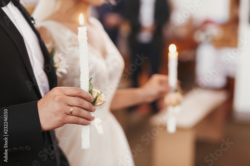 фотографія Bride and groom in golden crowns standing with candles in hands during wedding ceremony