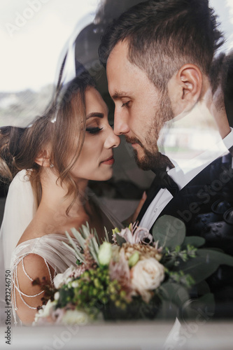 Gorgeous bride and stylish groom gently hugging at window. Sensual wedding couple embracing. Romantic moments of newlyweds. Creative wedding photo through glass. Copy space