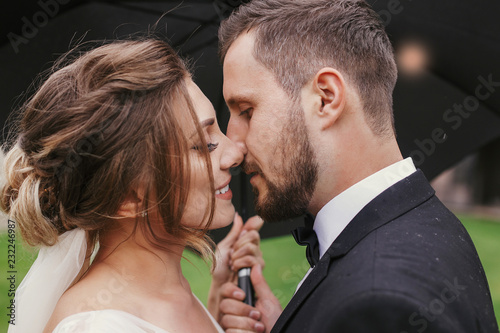 Gorgeous bride and stylish groom passionately kissing under umbrella in rainy outdoors. Sensual wedding couple embracing. Romantic moments of newlyweds. Modern wedding photo