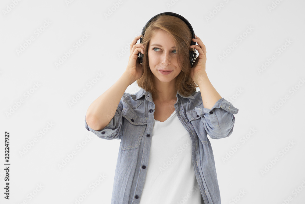 Music, relaxation and fun concept. Isolated picture of beautiful modern girl in blue shirt over white top looking away with pleased expression, enjoying good jazz tracks via wireless headphones Music, relaxation and fun concept. Isolated picture of beautiful modern girl in blue shirt over white top looking away with pleased expression, enjoying good jazz tracks via wireless headphones