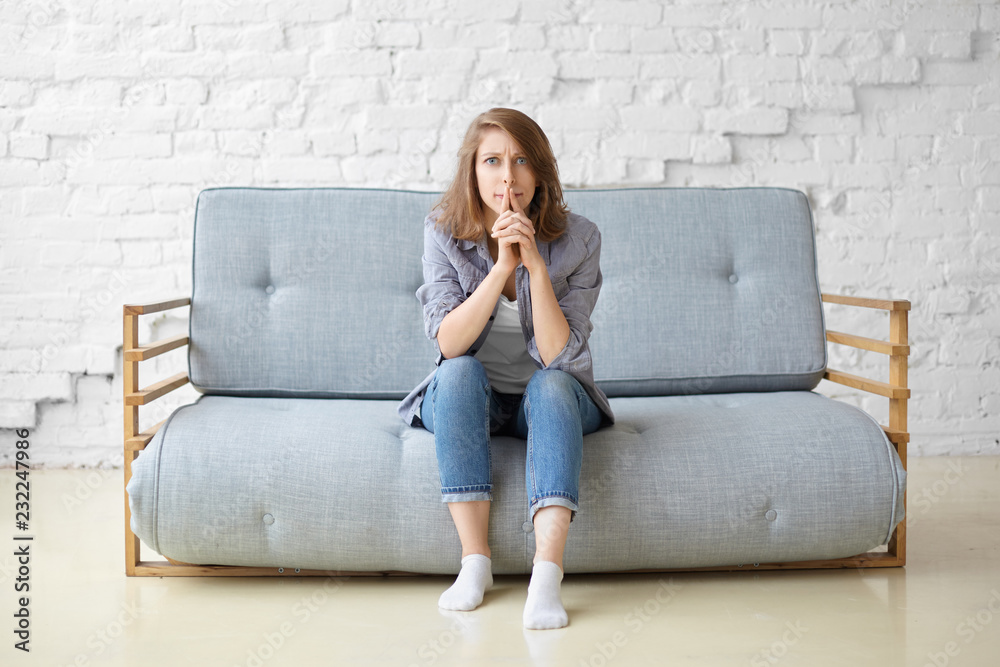 Concerned focused young woman in jeans and white socks sitting on grey couch against white brick wall background, having concentrated pensive look, pursing lips, thinking over home renovation ideas Concerned focused young woman in jeans and white socks sitting on grey couch against white brick wall background, having concentrated pensive look, pursing lips, thinking over home renovation ideas