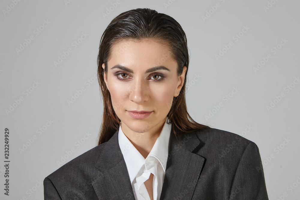 Close up image of trendy looking androgynal female with make up and hair combed back posing in studio, dressed in oversize gray men's jacket, looking at camera with serious facial expression Close up image of trendy looking androgynal female with make up and hair combed back posing in studio, dressed in oversize gray men's jacket, looking at camera with serious facial expression