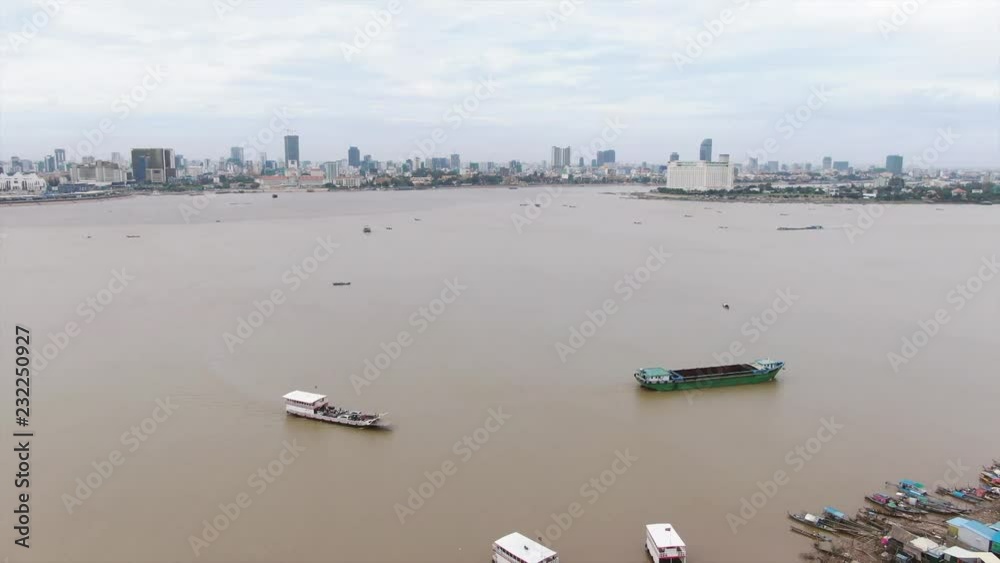 Aerial view of barge and ferry on Mekong river in Southeast Asia; camera moves back to reveal ferry landing with smaller boats and shops, with city in background. Aerial view of barge and ferry on Mekong river in Southeast Asia; camera moves back to reveal ferry landing with smaller boats and shops, with city in background.