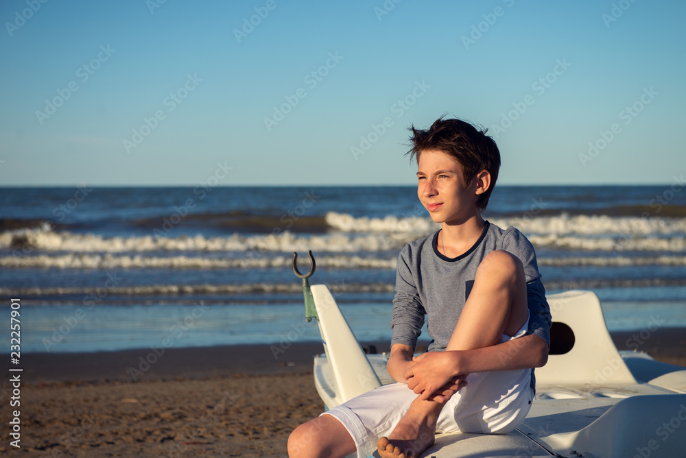 Young boy sitting on catamaran at the summer beach. Cute spectacled smiling happy 12 years old boy at seaside. Kid's outdoor portrait over seaside.