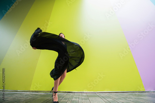 Fashion woman. Young beautiful chinese girl dancing outdoor wearing long black dress with high heels over colorful wall background. Stylish trendy lady. photo