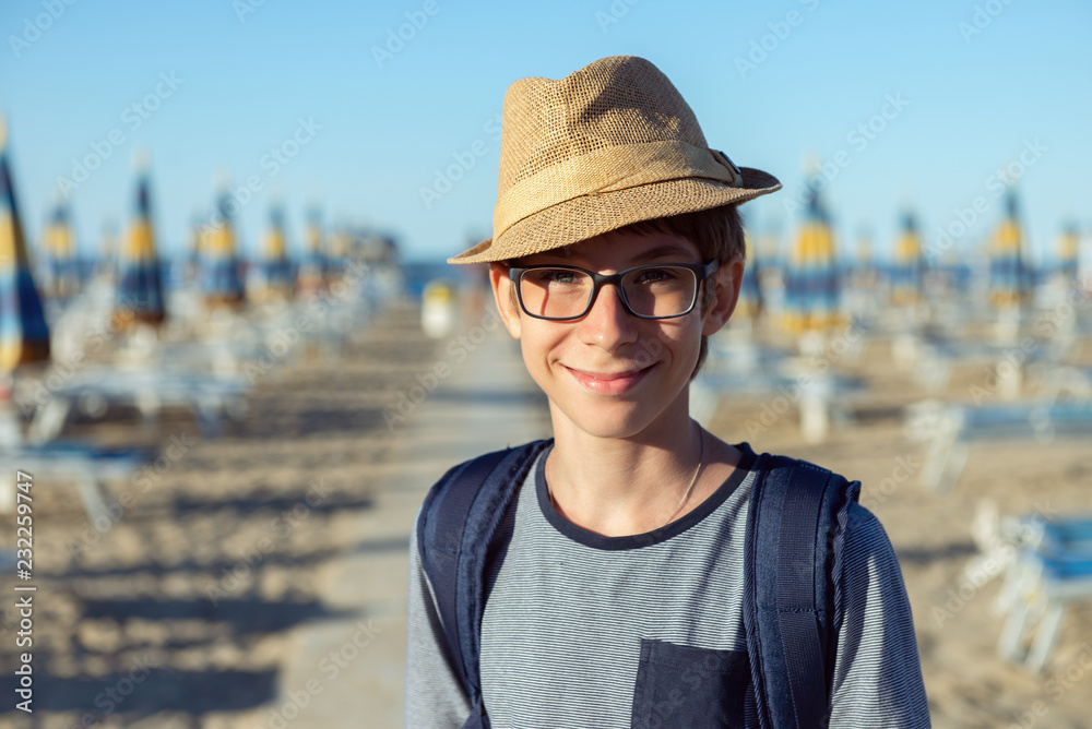 Young boy in hat and glasses posing at the summer beach. Cute smiling happy 12 years old boy at seaside, looking at camera. Kid's outdoor portrait over seaside.