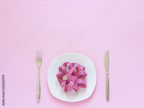 Photos Pink measuring tape lying on plate in the form of spaghetti, knife and fork on pink background