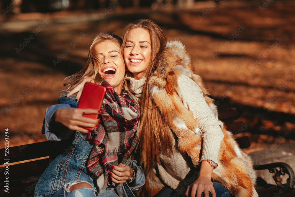 Fototapeta premium Two young women sitting on a bench and making selfie in the park