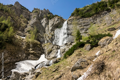 spring mountain waterfall, with green vegetation and snow on a blue sky 