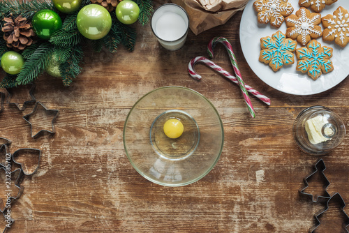 top view of raw chicken egg in bowl, christmas cookies on plate and decorative festive wreath on wooden surface