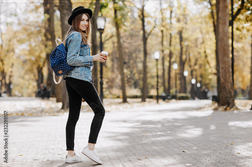 Image of a bright smiling hipster girl with brown hair wearing a hat, sunglasses and backpack holding cup of tea or coffee in the park © Screaghin