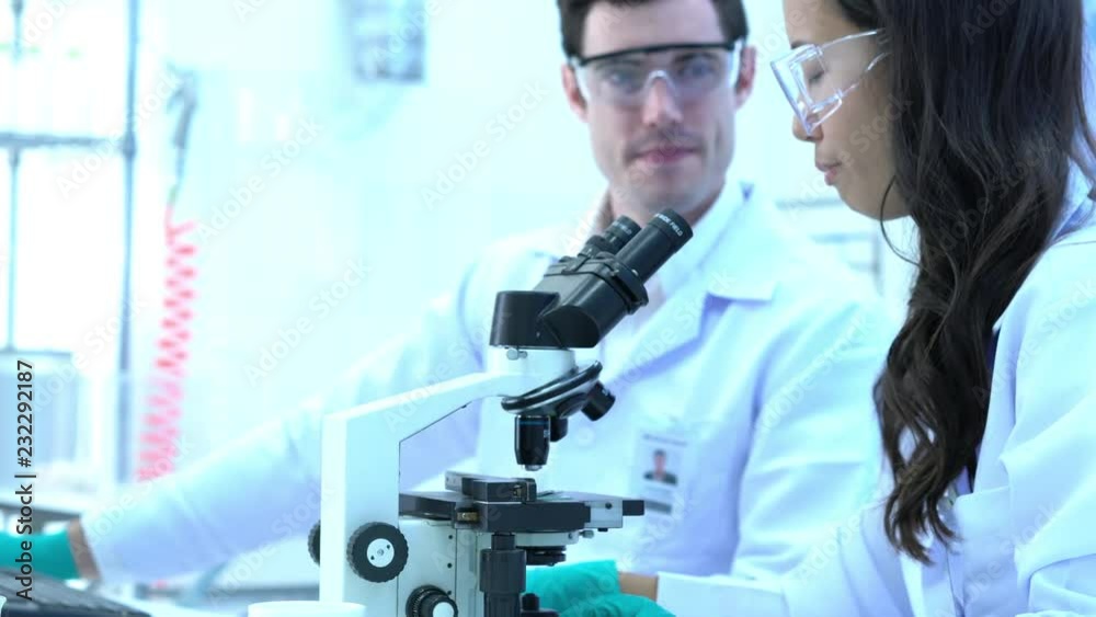 Scientist is looking through microscope with colleagues working in modern laboratory or medical center together. Concept of science, testing development and lab industry. Scientist is looking through microscope with colleagues working in modern laboratory or medical center together. Concept of science, testing development and lab industry.