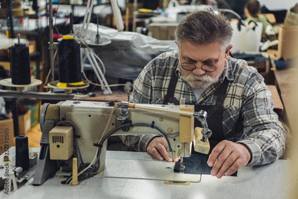 middle aged tailor in apron and eyeglasses working on sewing machine at studio middle aged tailor in apron and eyeglasses working on sewing machine at studio