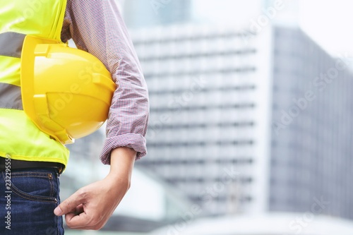 Close up front view of engineering male construction worker stand holding safety yellow helmet and wear reflective clothing for the safety of the work operation. outdoor of building background.