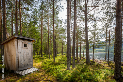 Autdoor wooden toilet in a beautiful sunny forest wilderness landscape by a lake.