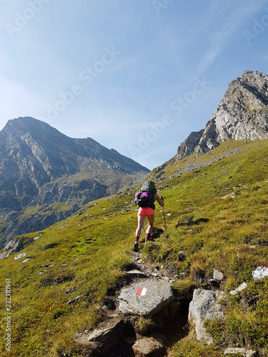 Hiker with backpack walking a grass trail on top of a mountain and enjoying valley view during trip in the alps
