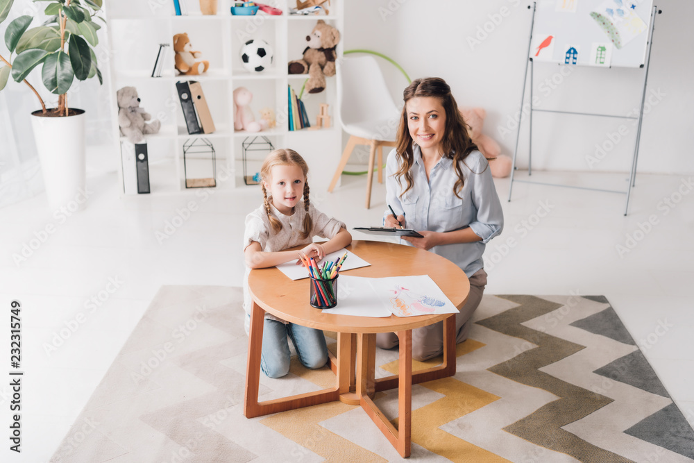 Obraz premium happy psychologist with clipboard sitting near little child while she drawing with color pencils and looking at camera